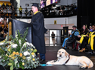 The shot is from stage left with Pat at the podium and guide dog Hogan attentively looking out at the crowd. Hogan is a yellow lab with an athletic build. Several people, wearing black gowns and caps and smiling, are seated behind Pat on stage. A portion of the crowd can be seen to the far left.
