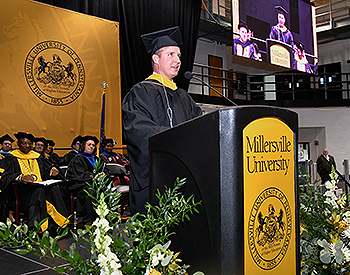 Taken from stage right, Pat delivers his speech. The stage party appears in the background to Pat&rsquo;s left. A large yellow and white flower arrangement is on both sides of the podium. Pat&rsquo;s image appears on a video screen at upper right of the photo.