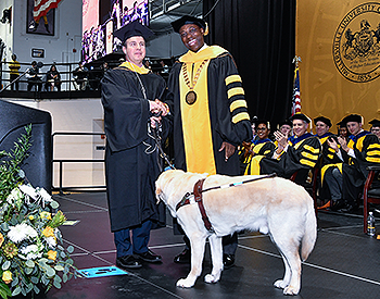 Pat shakes President Wubah&rsquo;s hand following the remarks. The seated stage party is clapping behind them. Guide Hogan stands in front of the pair, looking at Pat. The black and yellow MU seal can be seen behind the stage party.